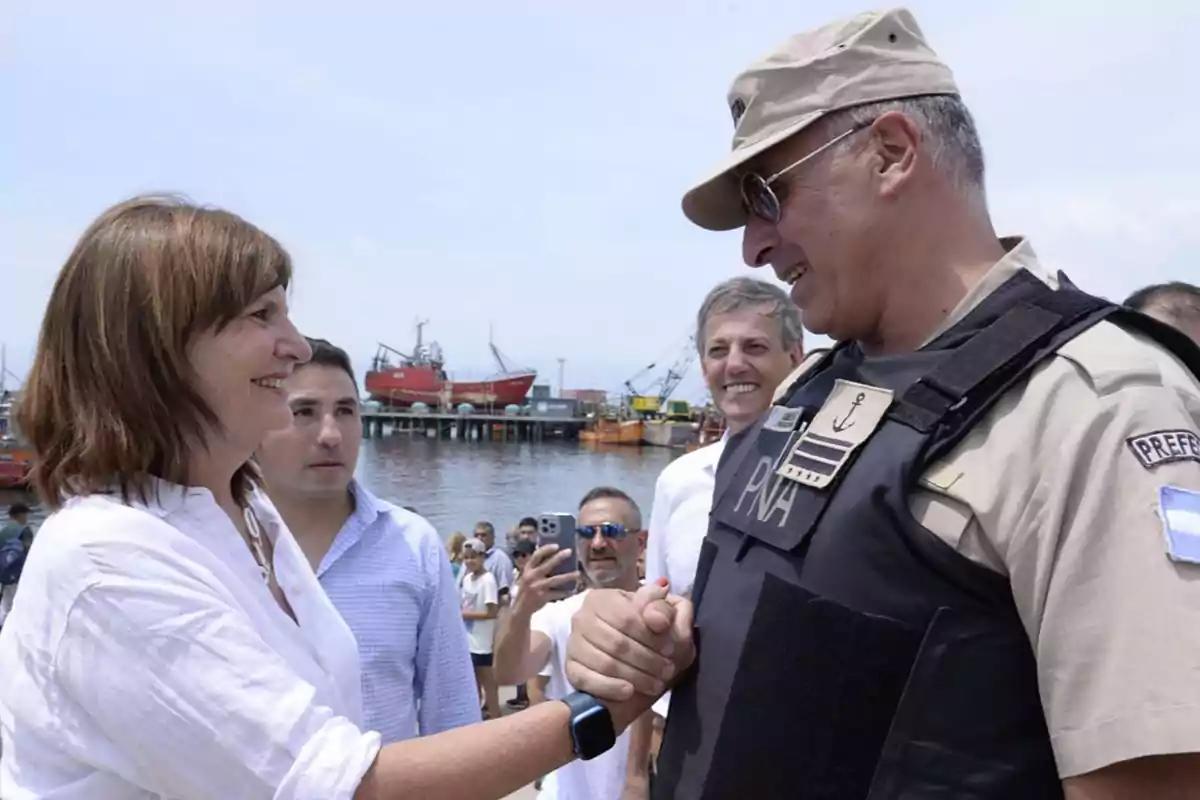 Una mujer sonríe y estrecha la mano de un hombre uniformado de la Prefectura Naval en un puerto mientras varias personas observan y barcos están de fondo Una mujer sonríe y estrecha la mano de un hombre uniformado de la Prefectura Naval en un puerto mientras varias personas observan y barcos están de fondo