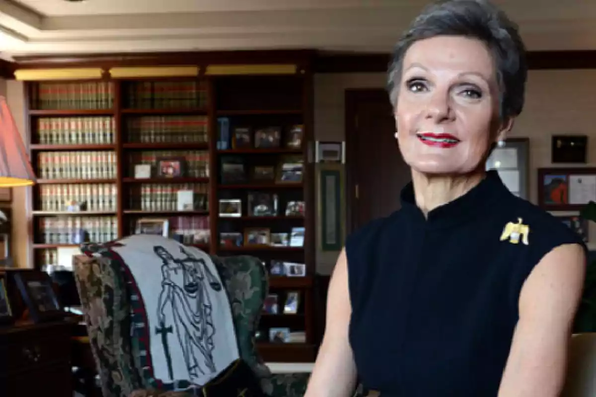Short-haired, gray-haired woman in a black dress sitting in an office with shelves full of books and photographs in the background Short-haired, gray-haired woman in a black dress sitting in an office with shelves full of books and photographs in the background