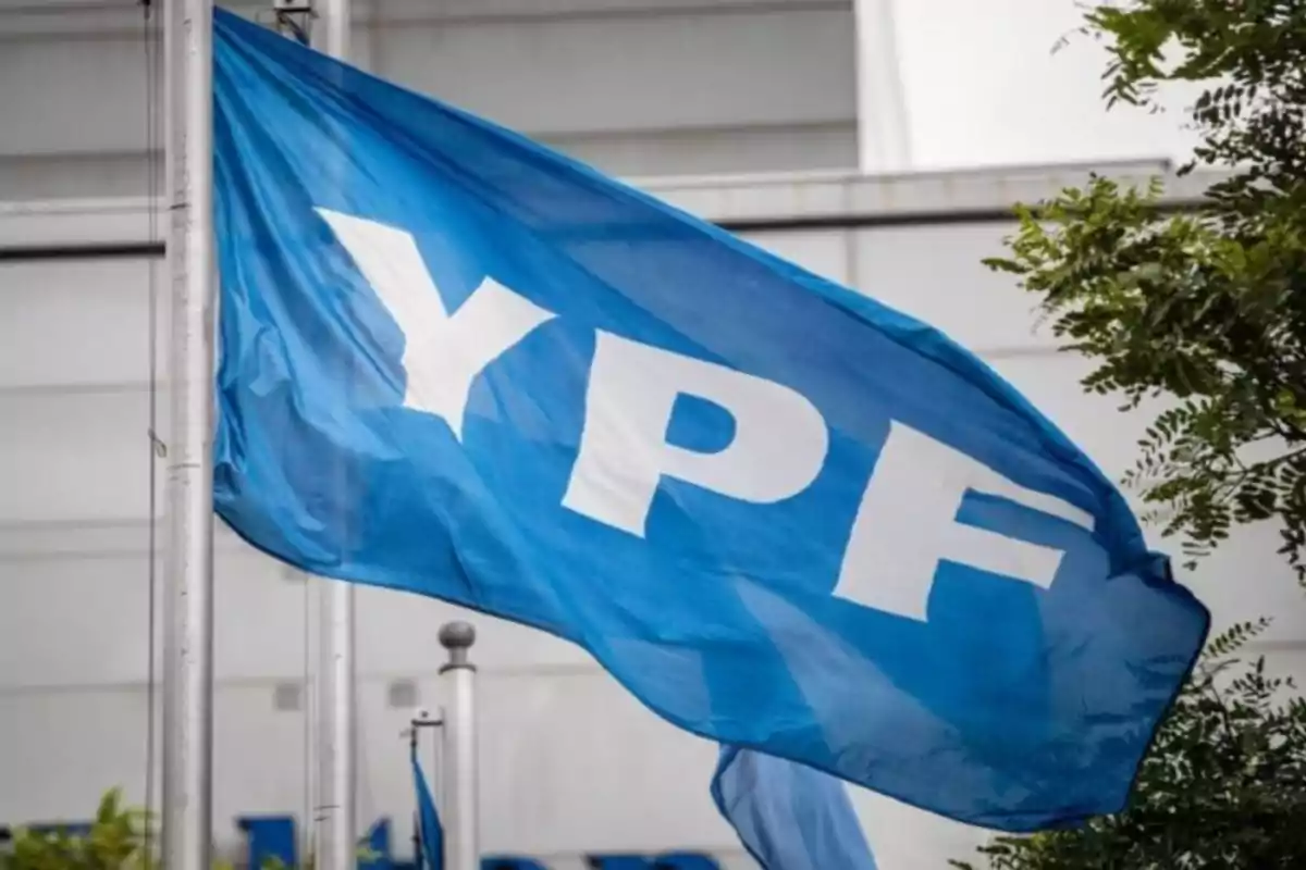 Bandera azul con las letras YPF en blanco ondeando al viento frente a un edificio y árboles Bandera azul con las letras YPF en blanco ondeando al viento frente a un edificio y árboles