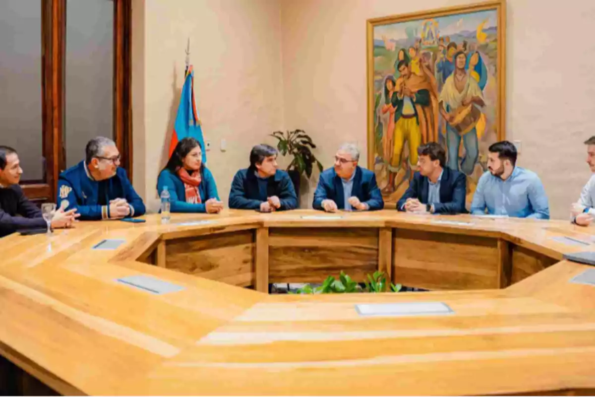 Eight people sitting around a wooden table in a meeting room with a flag and a colorful painting on the wall