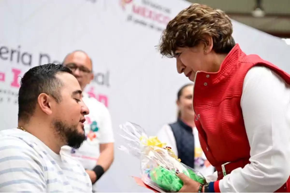 Una mujer con chaleco rojo entrega un regalo envuelto a un hombre con camiseta de rayas en un evento público