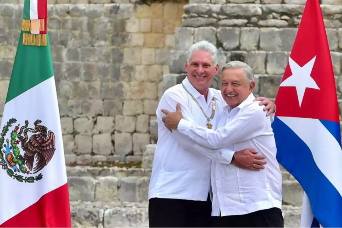 Two men smile and embrace in front of a stone structure, accompanied by the flags of Mexico and Cuba. Two men smile and embrace in front of a stone structure, accompanied by the flags of Mexico and Cuba.