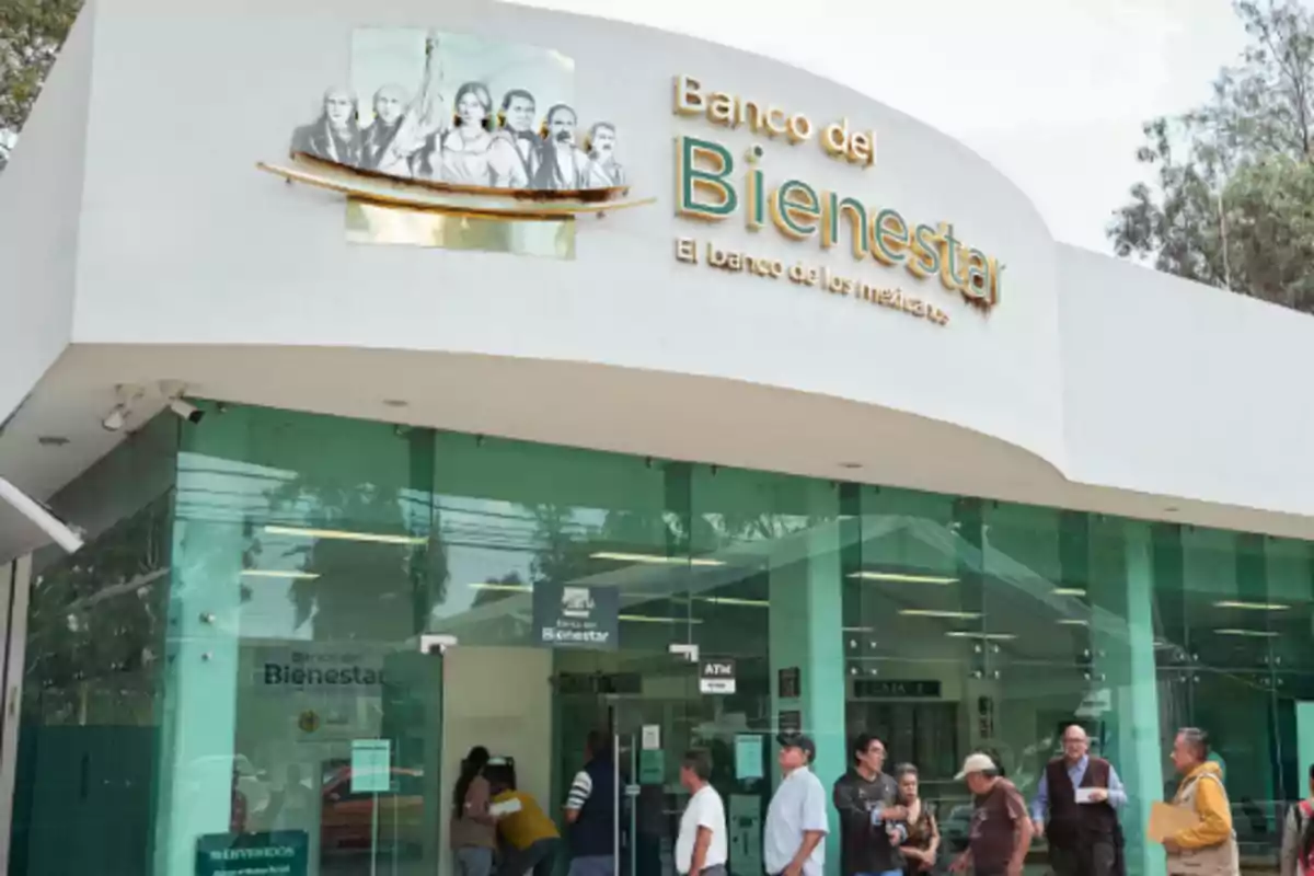 Personas formadas afuera de una sucursal del Banco del Bienestar con fachada de vidrio y letrero visible