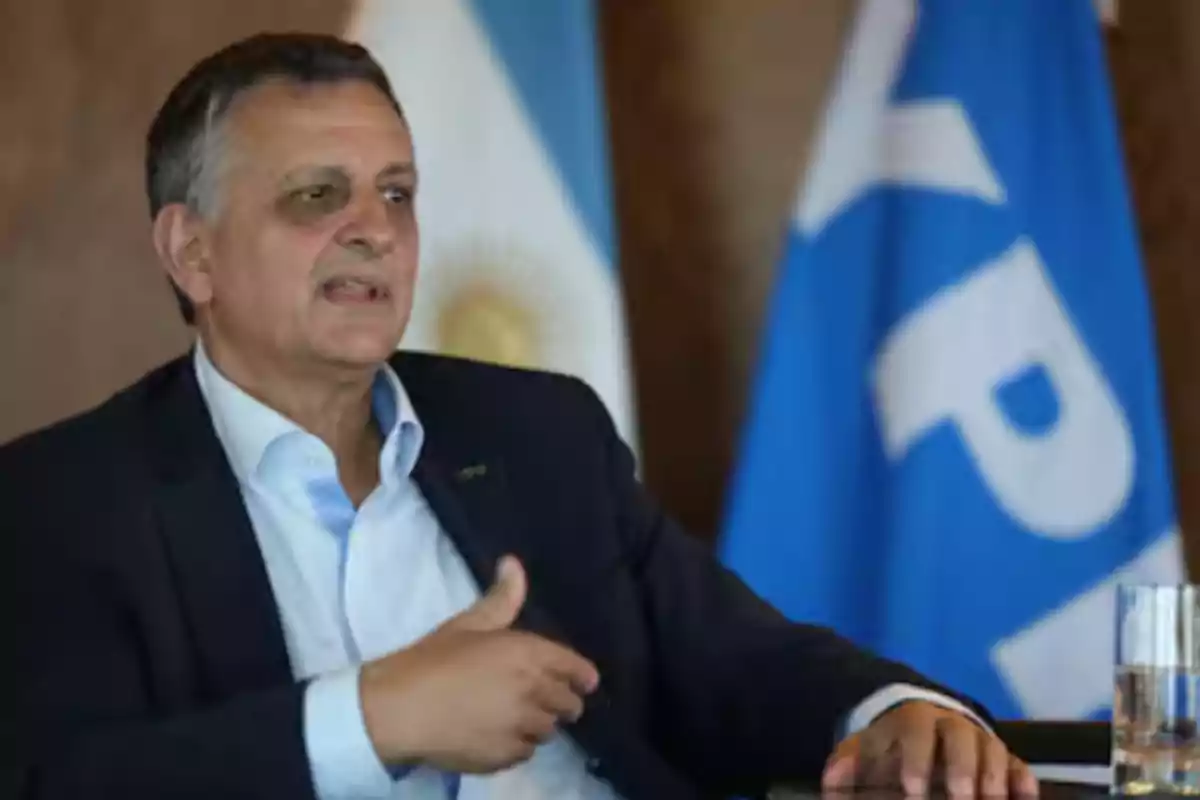 Hombre de mediana edad con traje oscuro sentado frente a banderas de Argentina y YPF mientras habla gesticulando con la mano Hombre de mediana edad con traje oscuro sentado frente a banderas de Argentina y YPF mientras habla gesticulando con la mano