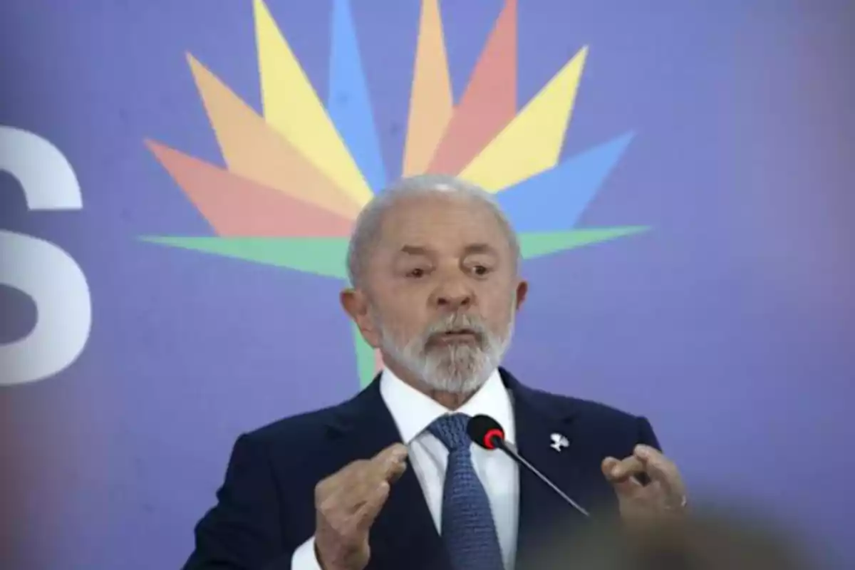 Gray-haired, bearded man speaking in front of a microphone with a blue background and a colorful design behind him Gray-haired, bearded man speaking in front of a microphone with a blue background and a colorful design behind him