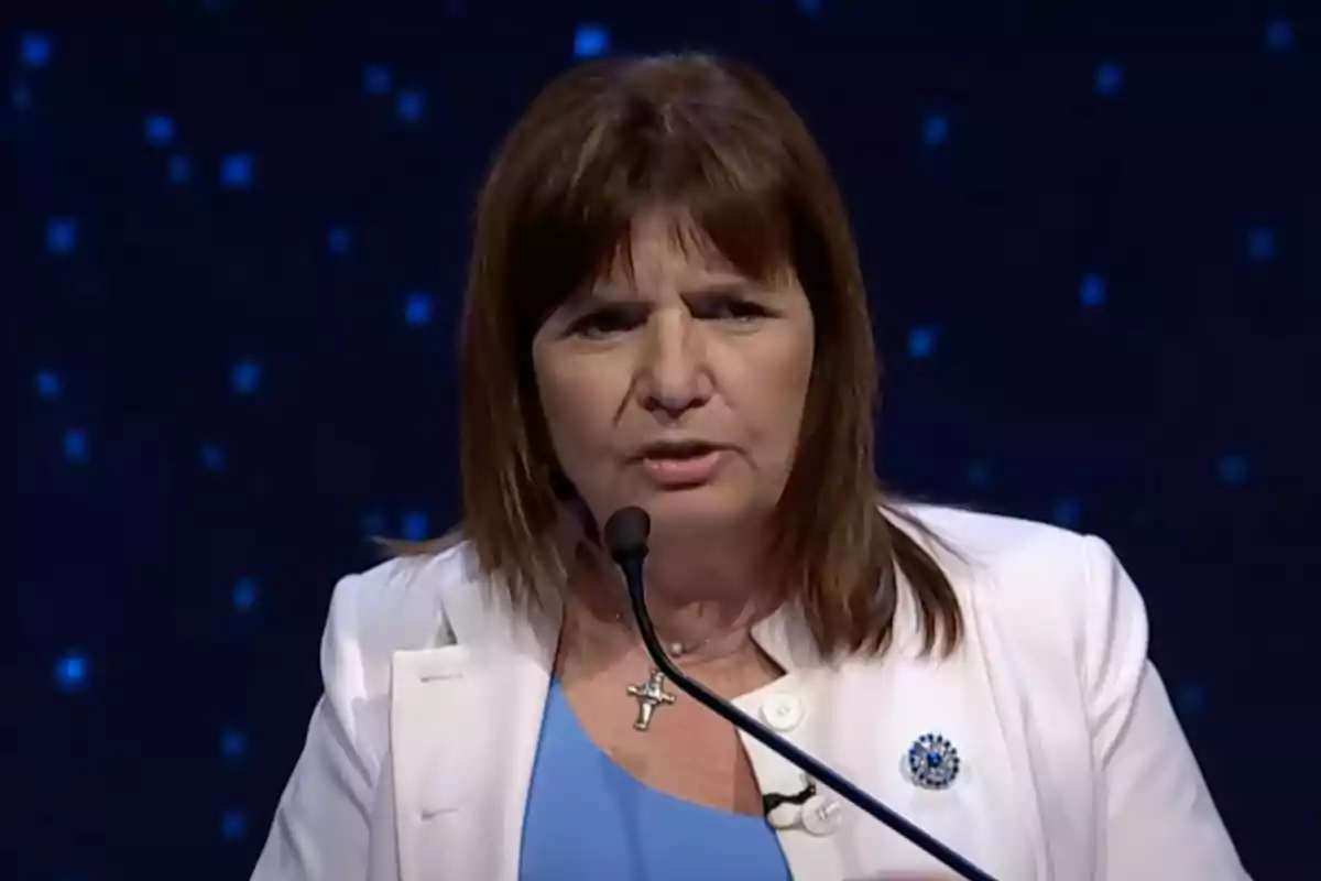 Brown-haired woman speaking in front of a microphone with a dark blue background and light spots, wearing a white blazer and light blue blouse