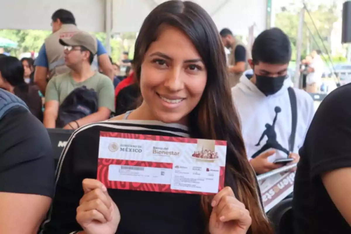 Smiling young person holds a Banco del Bienestar card from the Government of Mexico at an event with several people in the background. Smiling young person holds a Banco del Bienestar card from the Government of Mexico at an event with several people in the background.