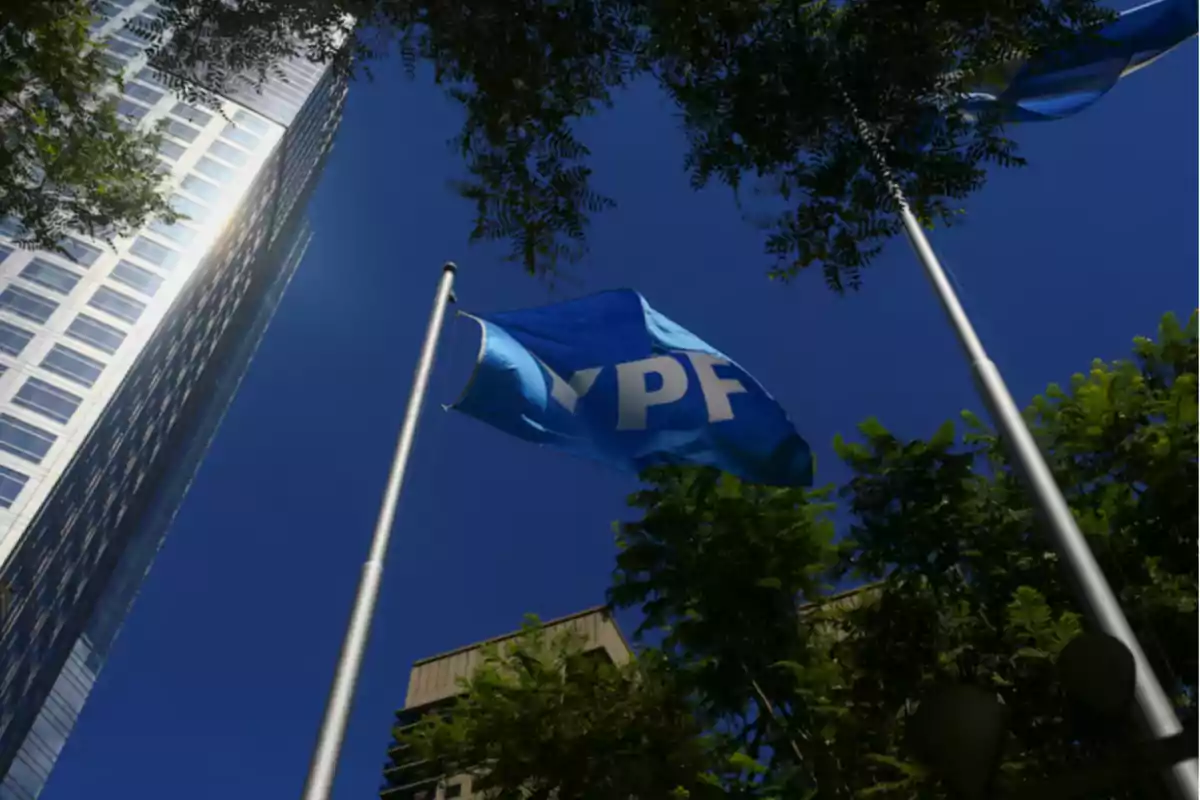 Bandera de YPF ondeando frente a un edificio alto rodeado de árboles bajo un cielo azul Bandera de YPF ondeando frente a un edificio alto rodeado de árboles bajo un cielo azul