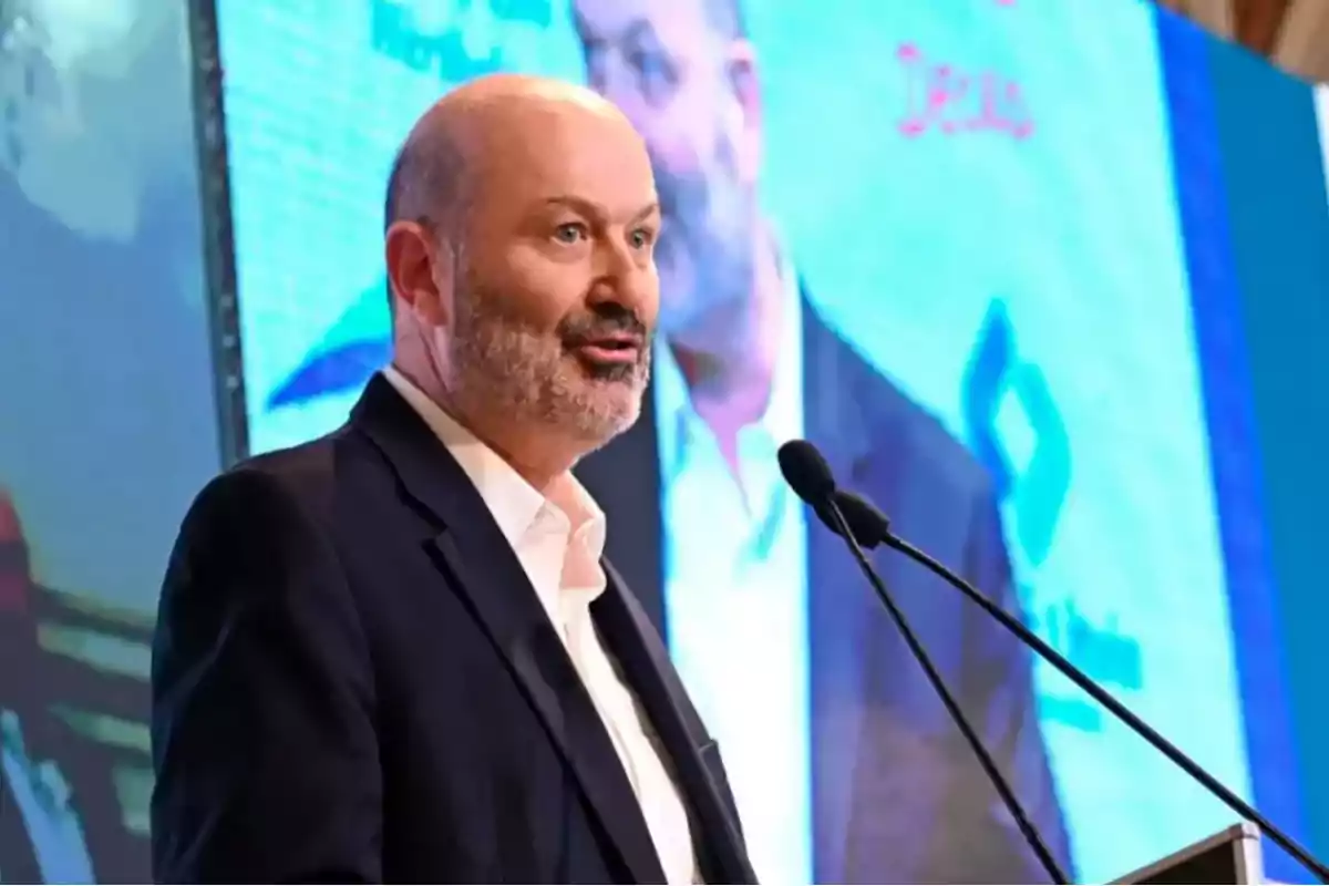Hombre calvo con barba hablando en un podio frente a un micrófono con una pantalla grande de fondo mostrando su imagen Hombre calvo con barba hablando en un podio frente a un micrófono con una pantalla grande de fondo mostrando su imagen