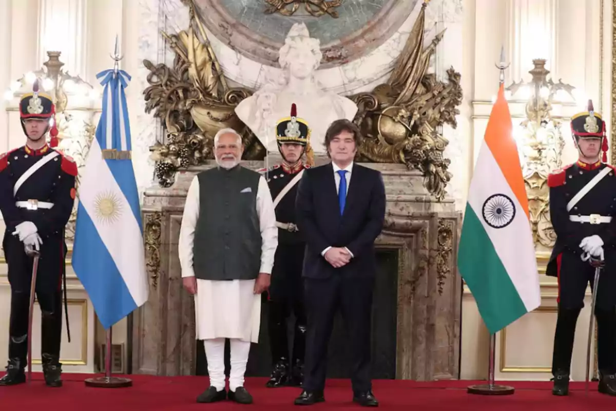 Dos hombres de pie en el centro de una sala elegante flanqueados por guardias con uniformes formales y las banderas de Argentina e India a los lados
