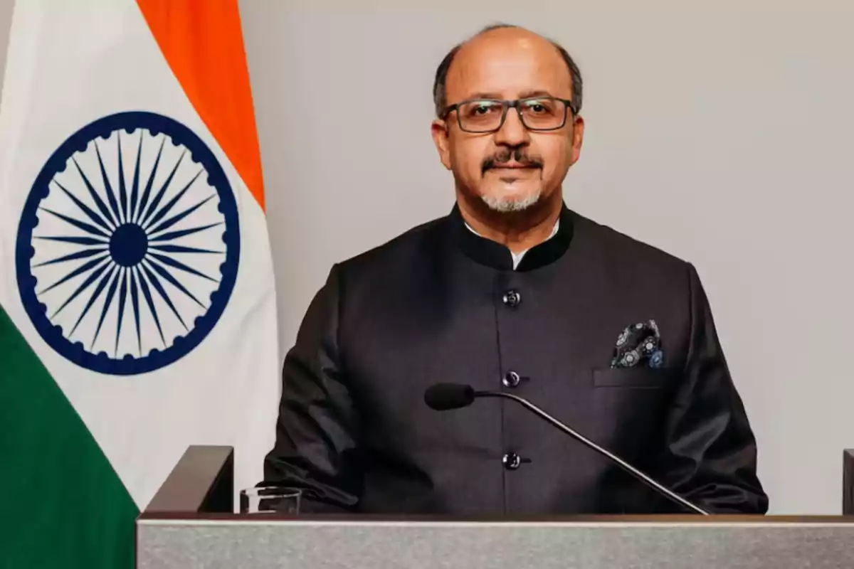 Hombre con gafas y barba de pie frente a un podio con un micrófono y la bandera de la India al fondo Hombre con gafas y barba de pie frente a un podio con un micrófono y la bandera de la India al fondo