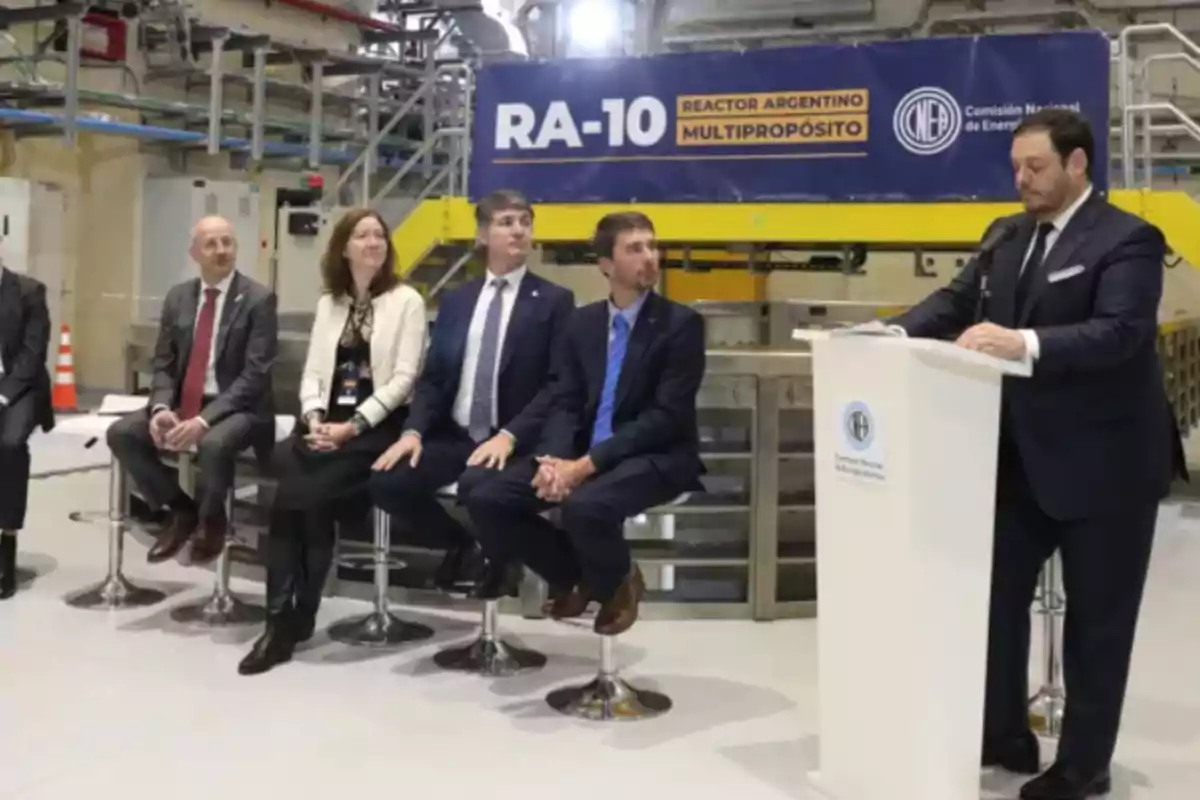 Un grupo de personas con vestimenta formal participa en un evento institucional frente a un cartel que dice RA-10 Reactor Argentino Multipropósito dentro de una instalación industrial Un grupo de personas con vestimenta formal participa en un evento institucional frente a un cartel que dice RA-10 Reactor Argentino Multipropósito dentro de una instalación industrial