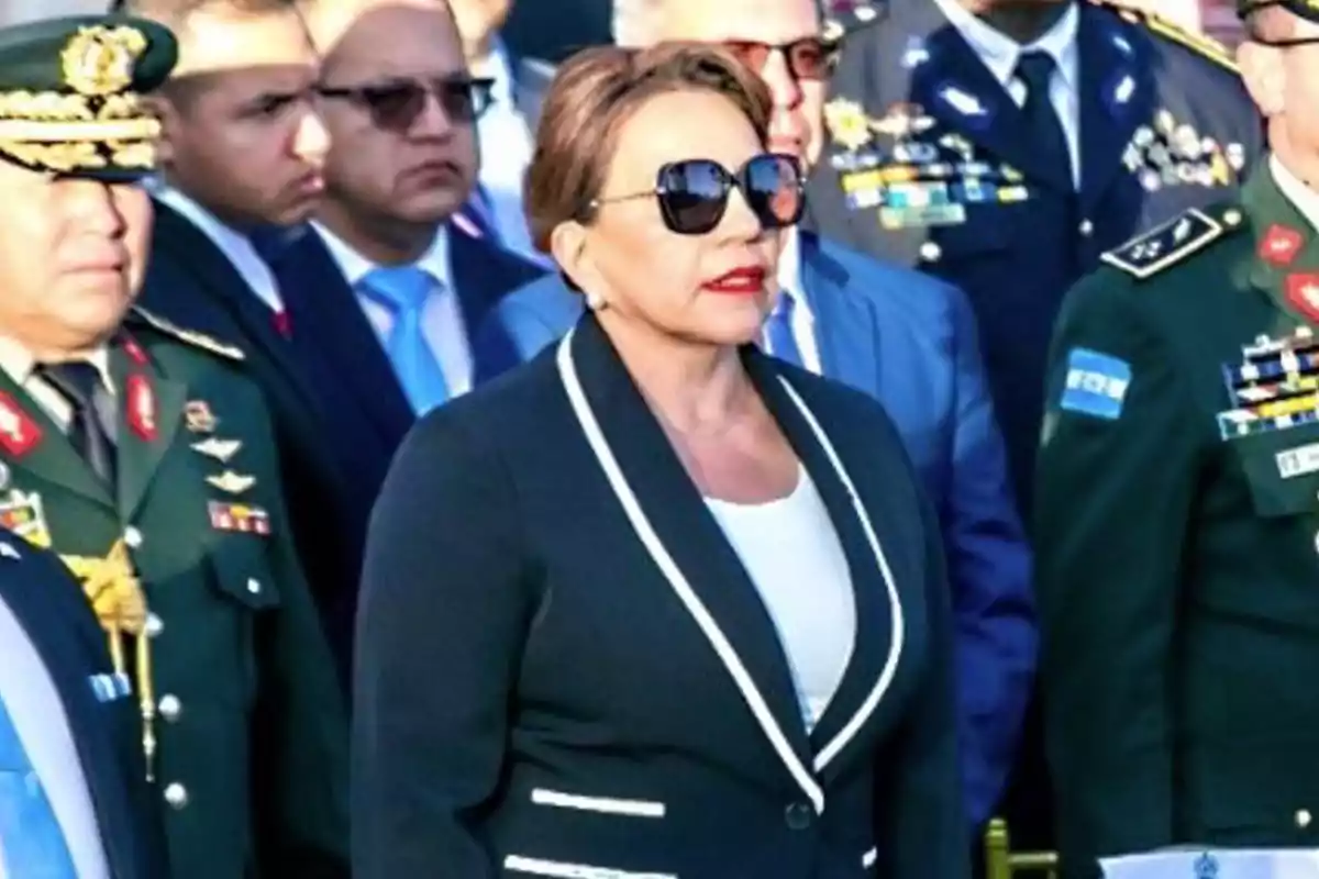 Mujer con gafas de sol y saco oscuro rodeada de hombres con uniformes militares y trajes formales en un evento oficial Mujer con gafas de sol y saco oscuro rodeada de hombres con uniformes militares y trajes formales en un evento oficial