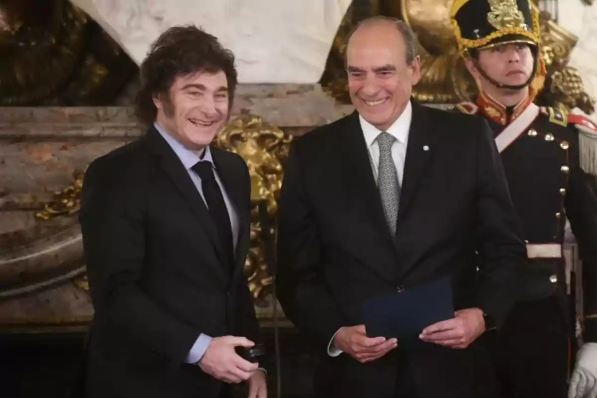 Two men in dark suits smile as they pose together in a formal setting with golden decor and a guard in ceremonial uniform in the background.