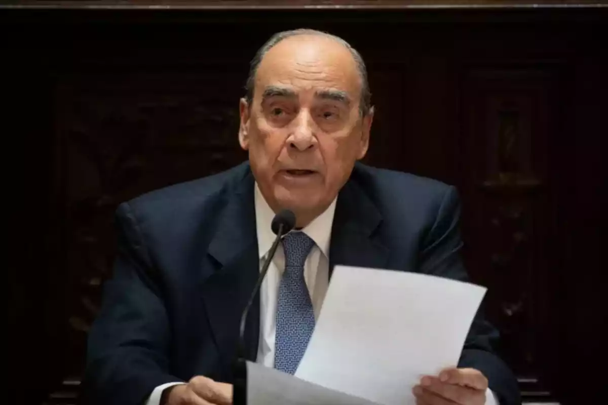 Older man in a dark suit speaking in front of a microphone while holding some papers in his hands Older man in a dark suit speaking in front of a microphone while holding some papers in his hands