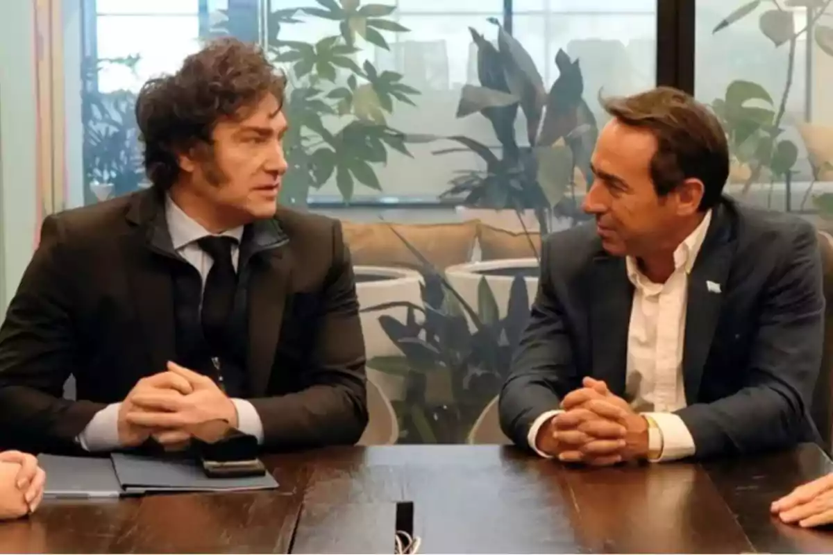 Two men dressed in suits are talking while sitting at a wooden table in an office with plants in the background. Two men dressed in suits are talking while sitting at a wooden table in an office with plants in the background.