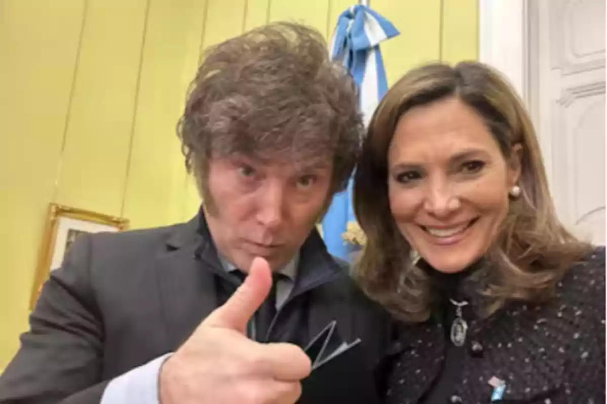 Two people pose for a selfie, one of them is smiling and the other is giving a thumbs-up, with a light blue and white flag visible in the background. Two people pose for a selfie, one of them is smiling and the other is giving a thumbs-up, with a light blue and white flag visible in the background.