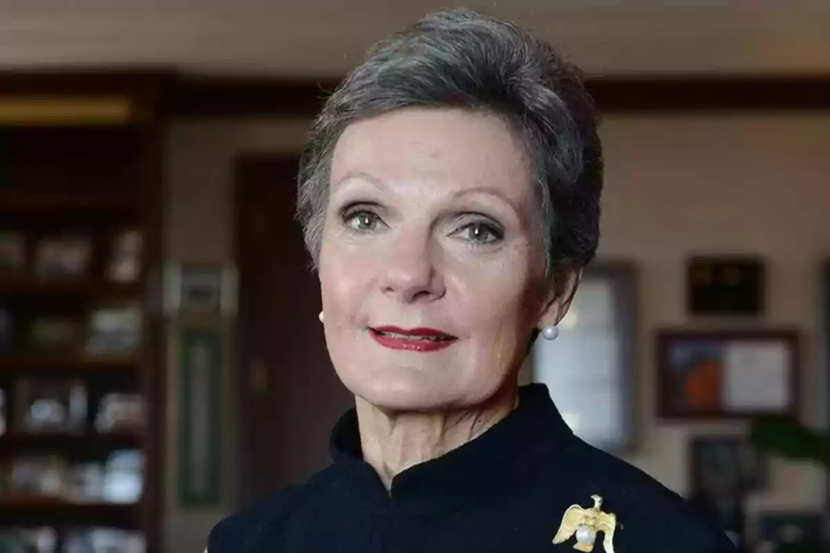 Short-haired, gray-haired woman with a gold eagle-shaped brooch on her dark jacket, posing in an elegant indoor setting. Short-haired, gray-haired woman with a gold eagle-shaped brooch on her dark jacket, posing in an elegant indoor setting.