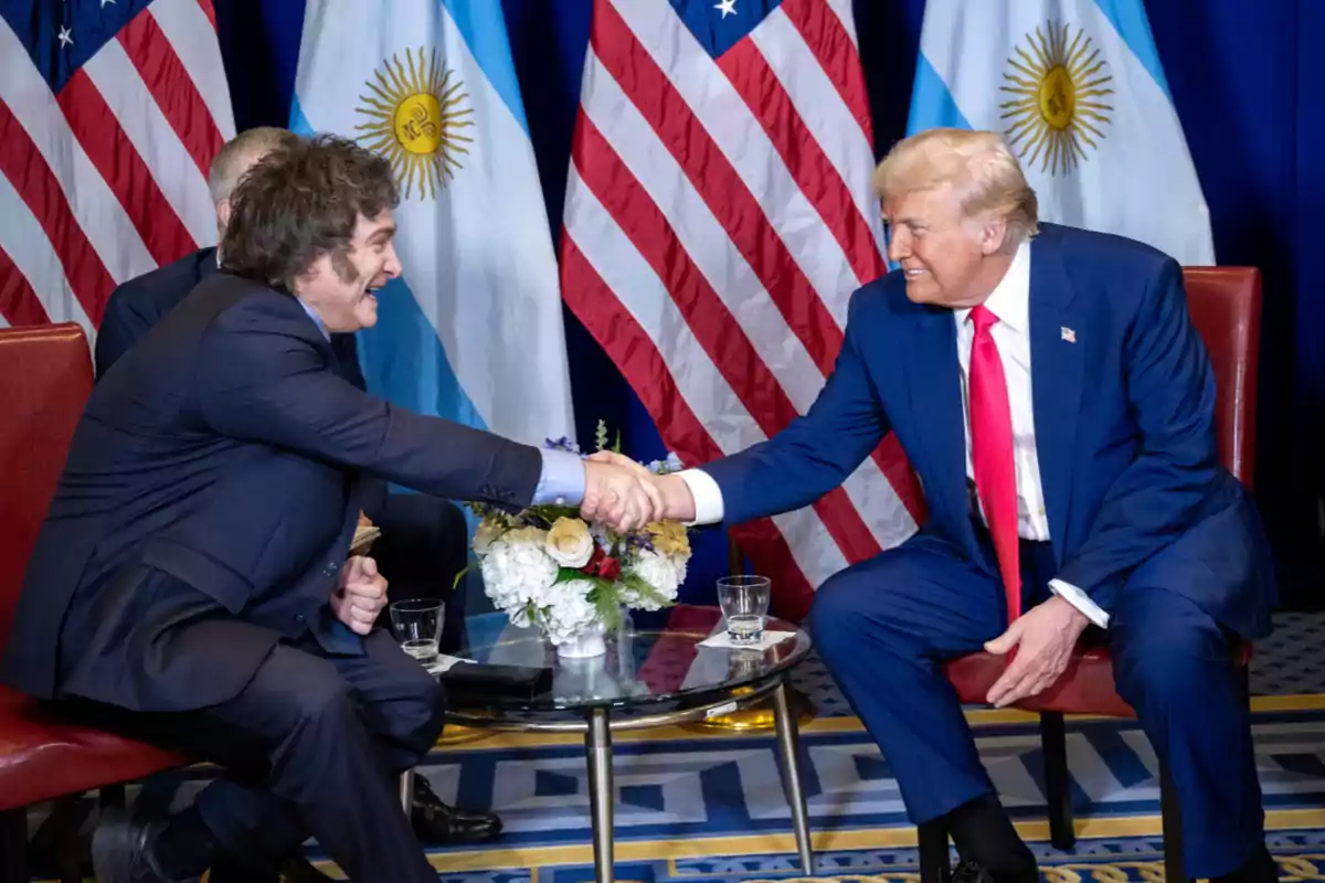 Two men in suits shake hands while sitting face to face in a room with Argentine and United States flags in the background.