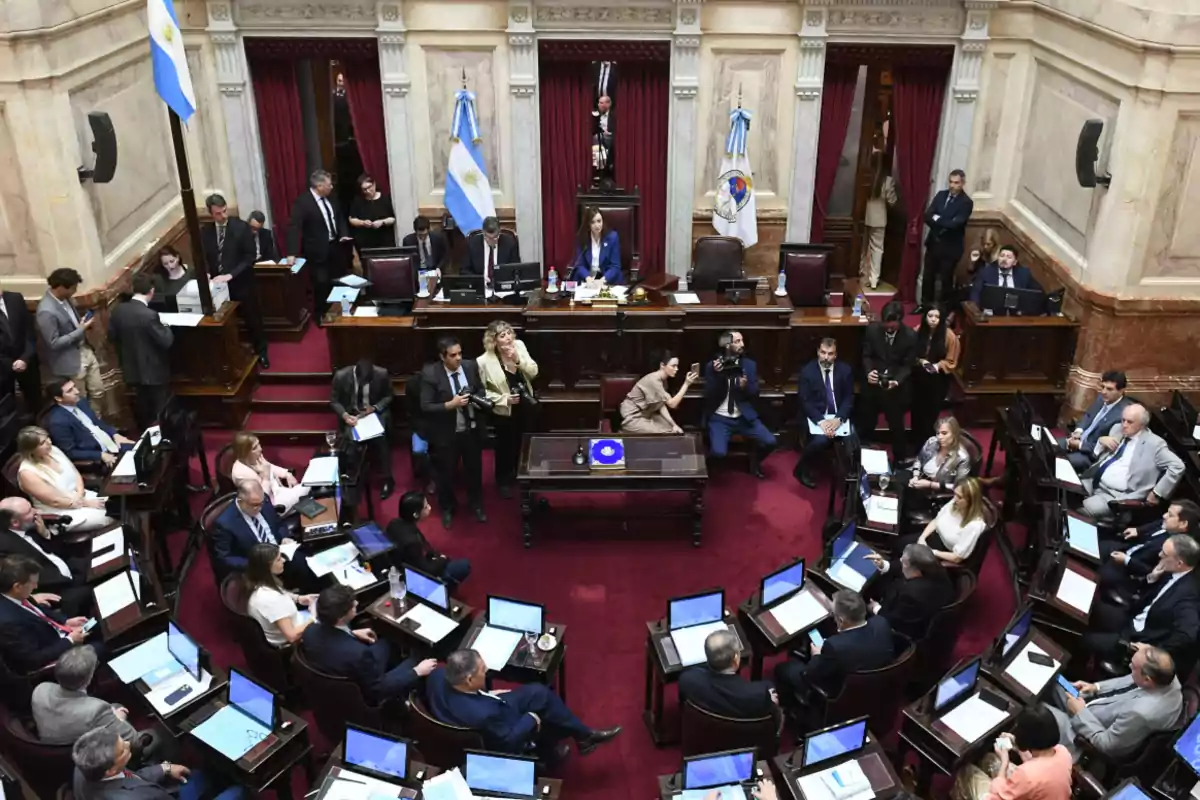 Overview of the Argentine Senate chamber during a session, with senators seated at their desks, laptops open, and several people standing, including photographers and staff, in a formal and solemn atmosphere. Overview of the Argentine Senate chamber during a session, with senators seated at their desks, laptops open, and several people standing, including photographers and staff, in a formal and solemn atmosphere.