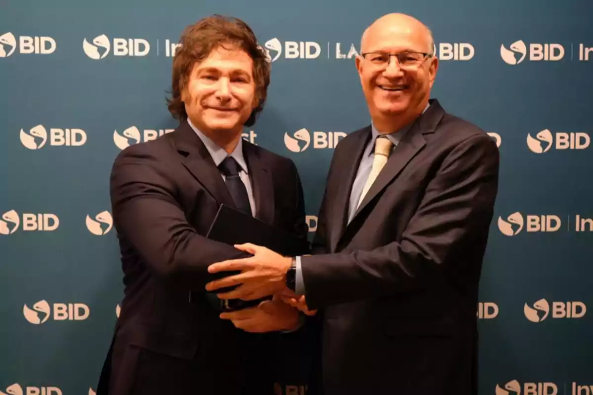 Two men in suits shake hands and smile in front of a blue background with the IDB logo.