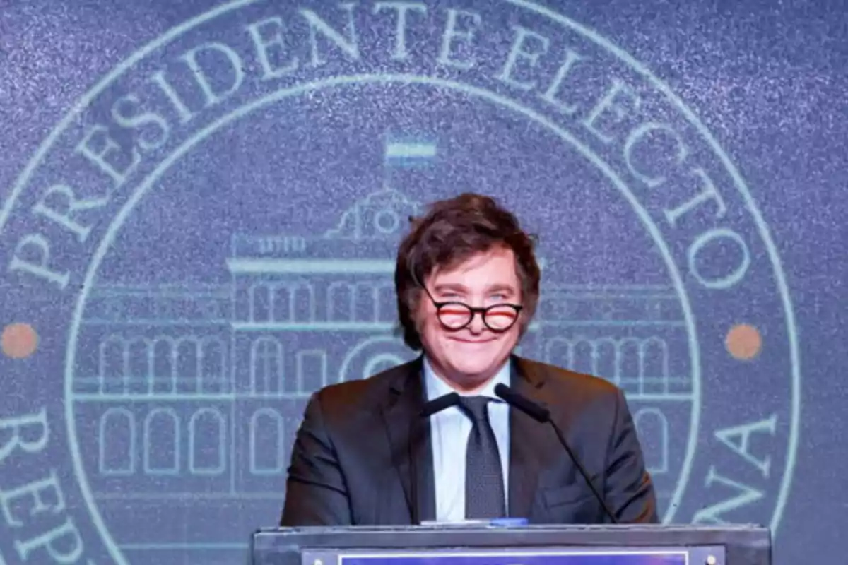 Dark-haired man with round-framed glasses speaking at a podium with a blue background featuring a presidential seal and the words Dark-haired man with round-framed glasses speaking at a podium with a blue background featuring a presidential seal and the words