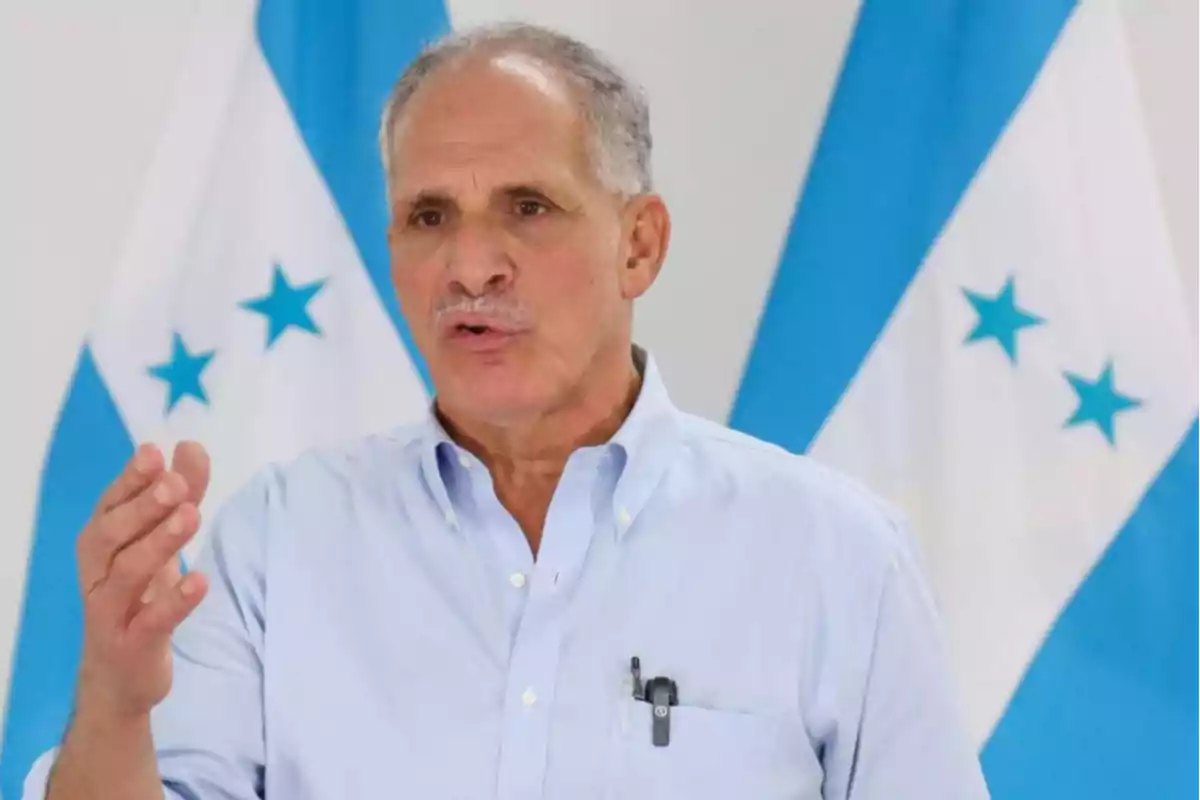 Gray-haired man with a mustache speaking with gestures in front of two Honduran flags