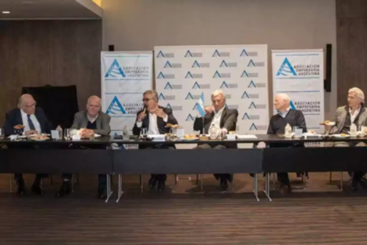 Seis hombres de traje sentados en una mesa larga durante una reunión empresarial con carteles de la Asociación Empresaria Argentina de fondo
