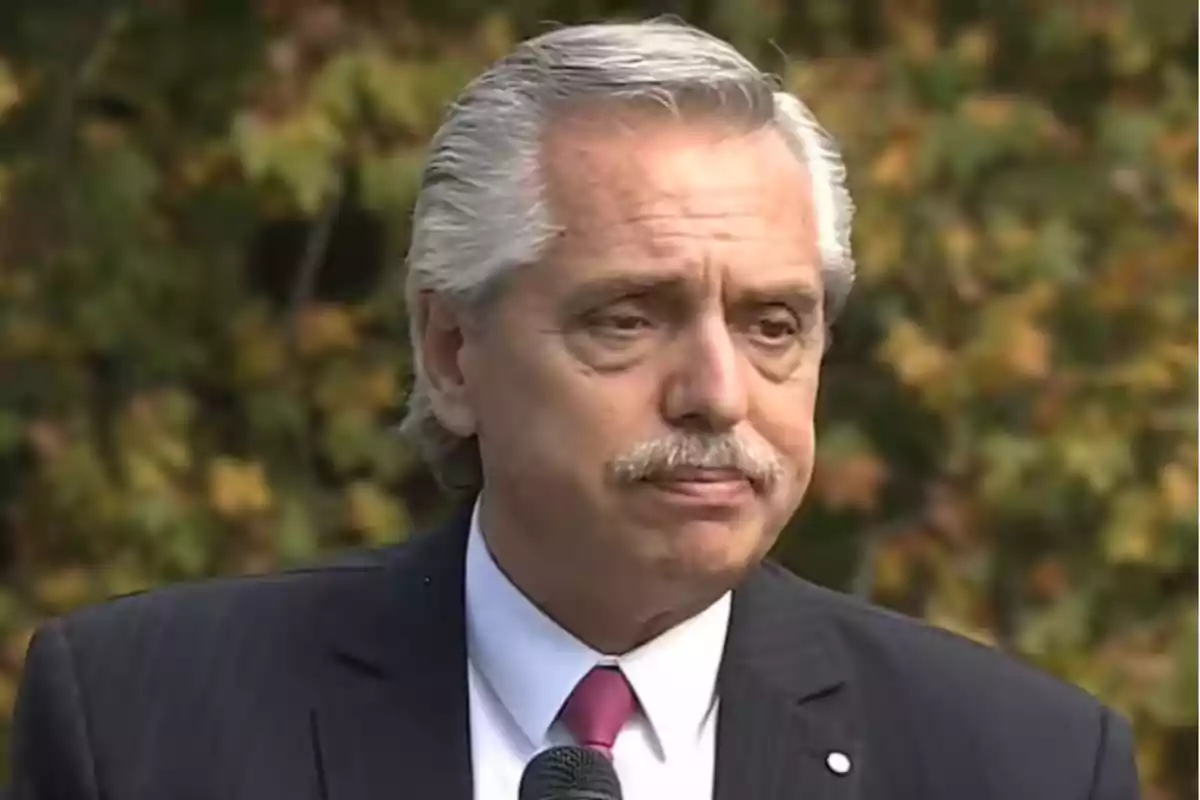 Hombre de cabello canoso y bigote con expresión seria usando traje y corbata frente a un micrófono y un fondo de árboles.