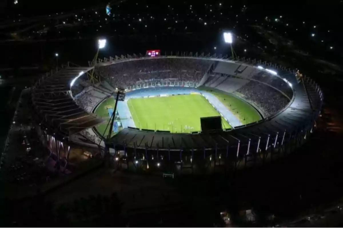 Vista aérea de un estadio de fútbol iluminado durante la noche con las gradas llenas de espectadores y el campo de juego en el centro