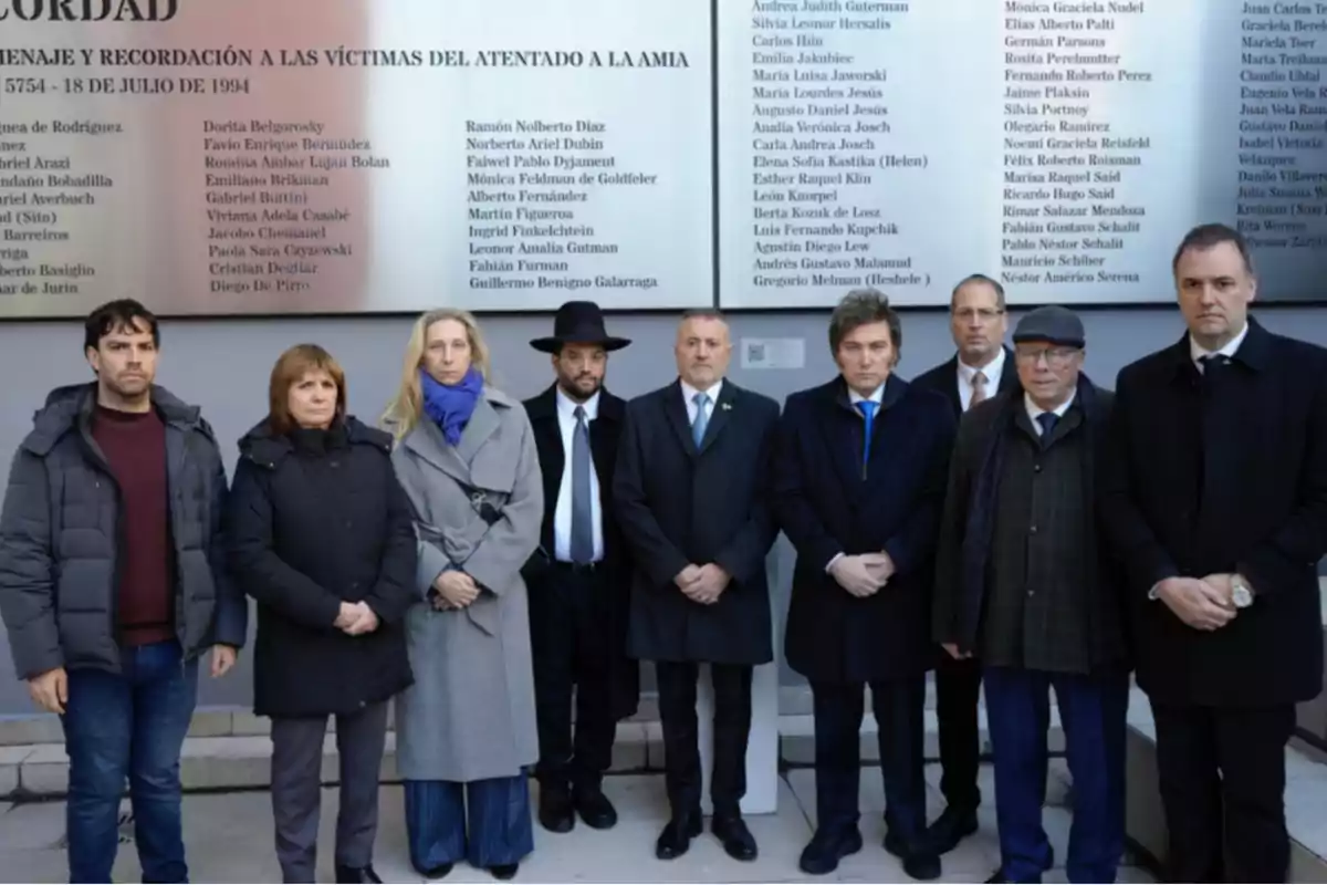 Nueve personas posan de pie frente a una placa conmemorativa con nombres, en un acto de homenaje y recordación a las víctimas del atentado a la AMIA.