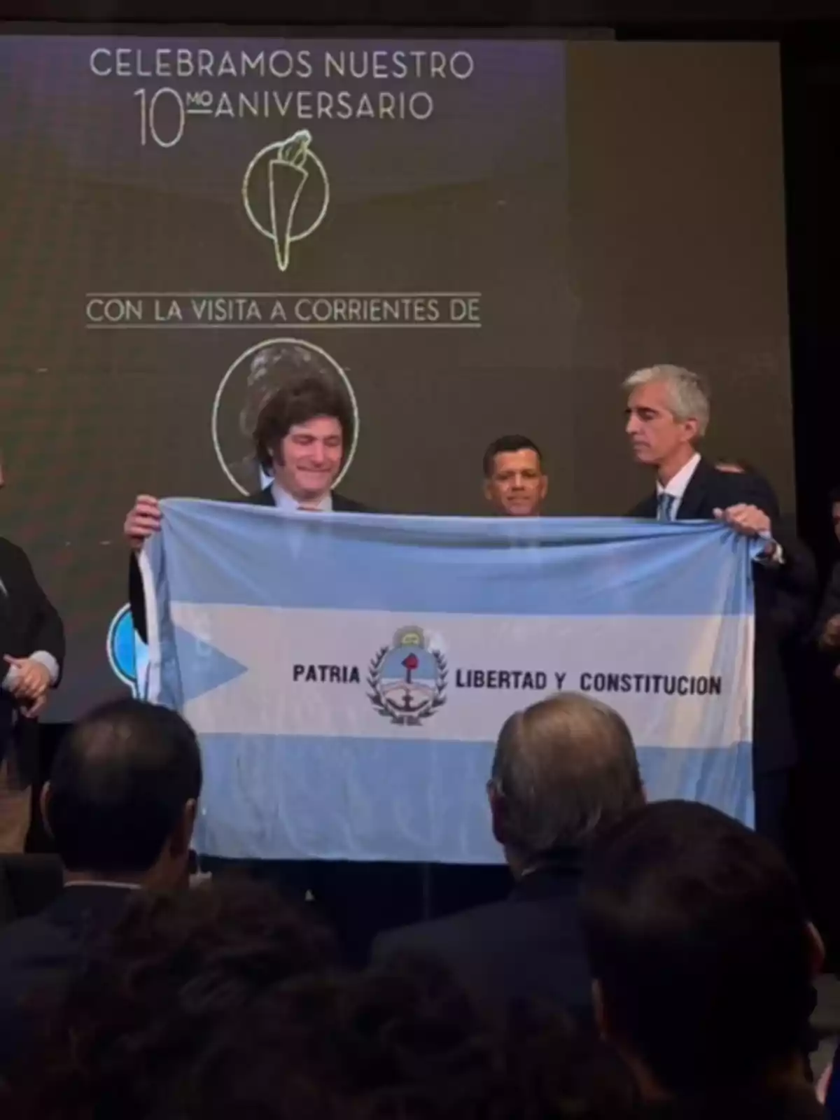Un hombre sonriente sostiene una bandera celeste y blanca con la inscripción Patria Libertad y Constitución junto a otras personas en un evento con un cartel de aniversario de fondo