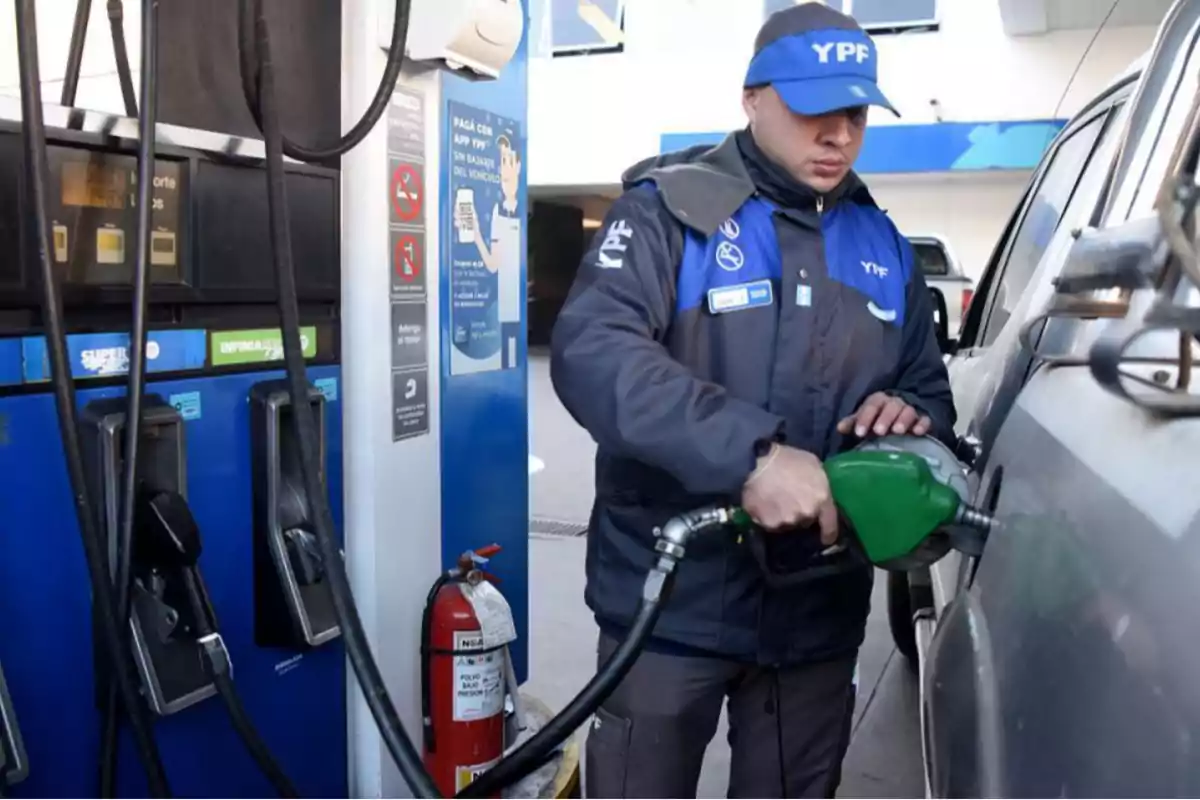 Empleado de estación de servicio con uniforme azul cargando combustible en un auto junto a un surtidor y un extintor