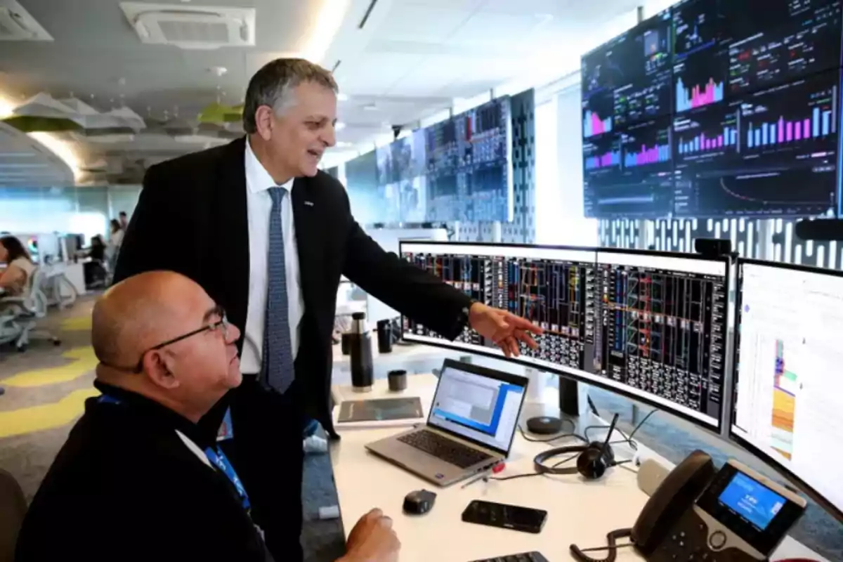 Two people in a modern office observe several screens with charts and financial data while they talk and point at the information on the monitors. Two people in a modern office observe several screens with charts and financial data while they talk and point at the information on the monitors.