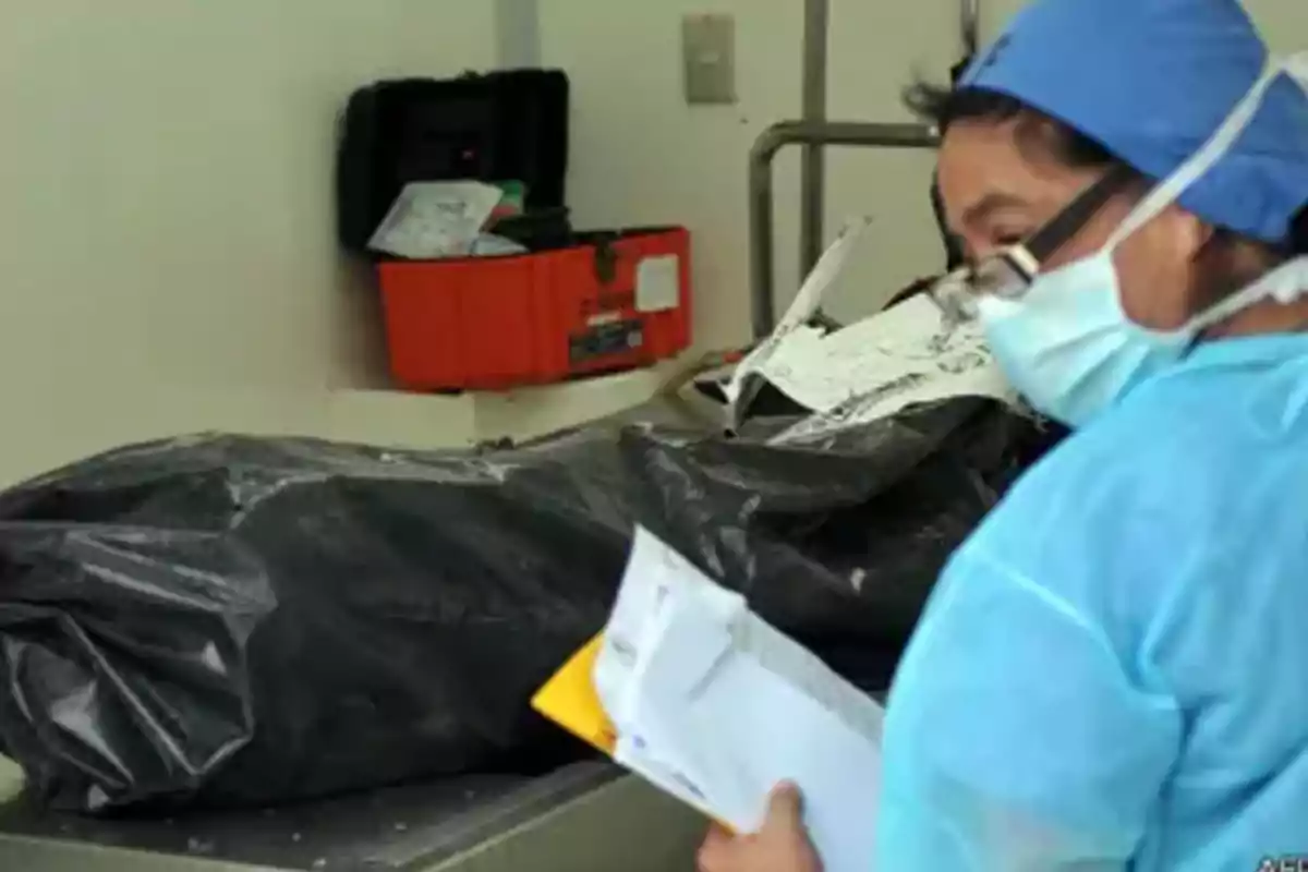 Persona con bata y cubrebocas revisando documentos junto a una bolsa negra sobre una camilla en una sala médica
