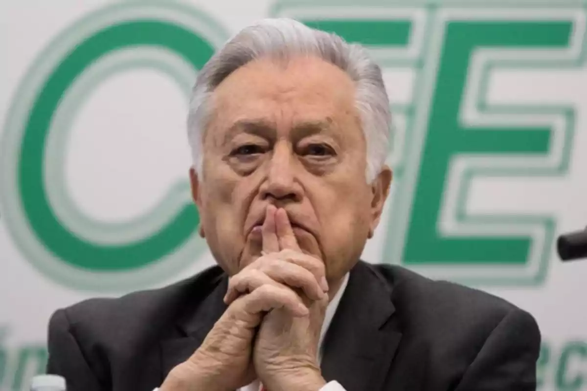 Hombre mayor de cabello canoso con expresión seria y manos entrelazadas frente a su rostro, al fondo letras verdes desenfocadas