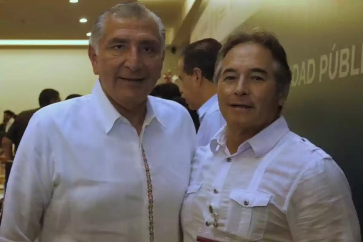 Two men pose together at a formal event, both wearing white jerseys and standing indoors with other people in the background. Two men pose together at a formal event, both wearing white jerseys and standing indoors with other people in the background.