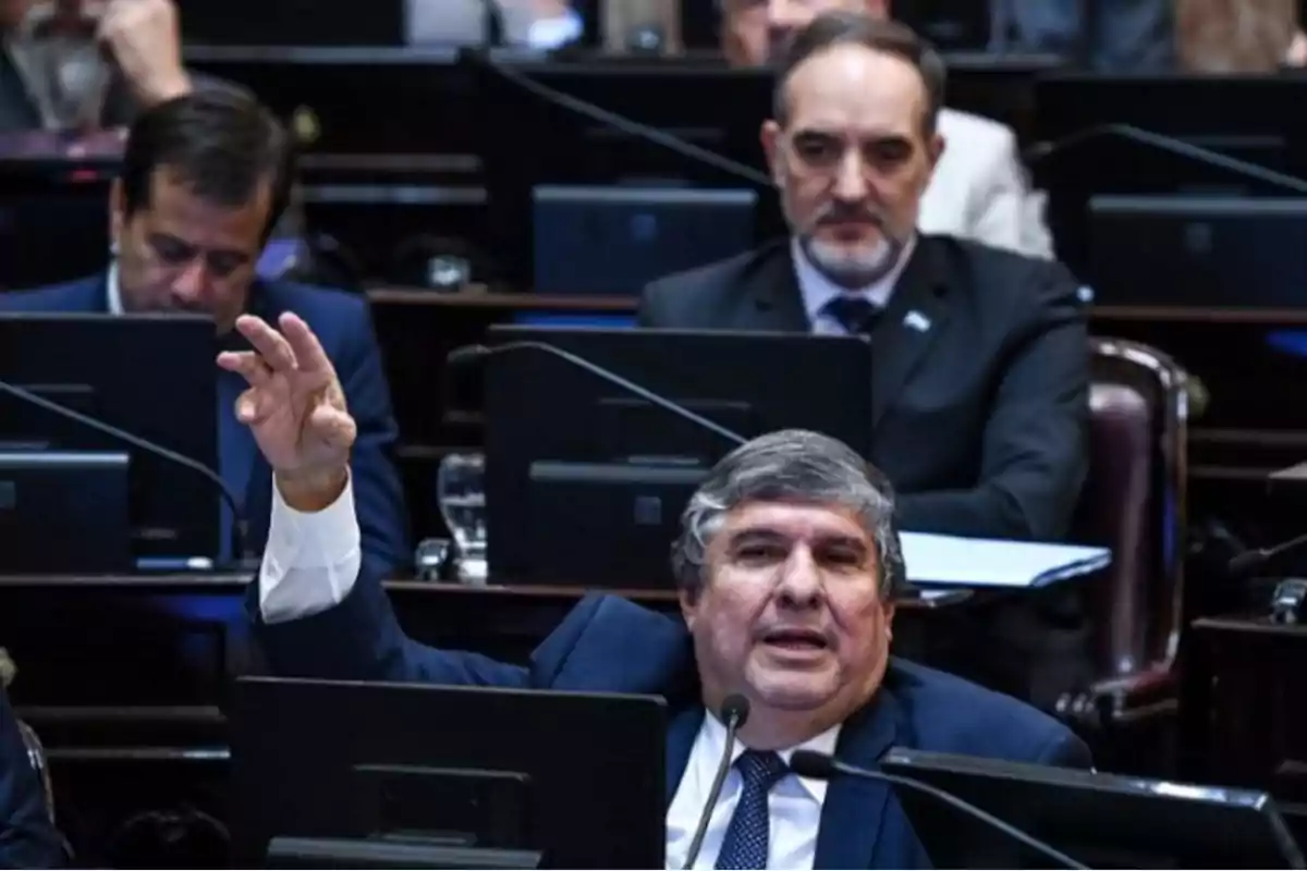Un hombre de traje y corbata habla y gesticula con la mano levantada en una sala de sesiones parlamentarias, mientras otras personas lo observan o trabajan en sus computadoras.