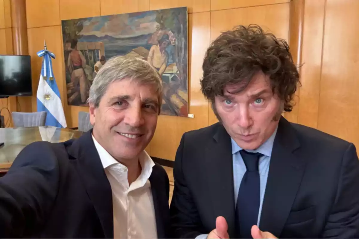 Dos hombres vestidos de traje posan para una selfie en una oficina con una bandera de Argentina y un cuadro en la pared de fondo