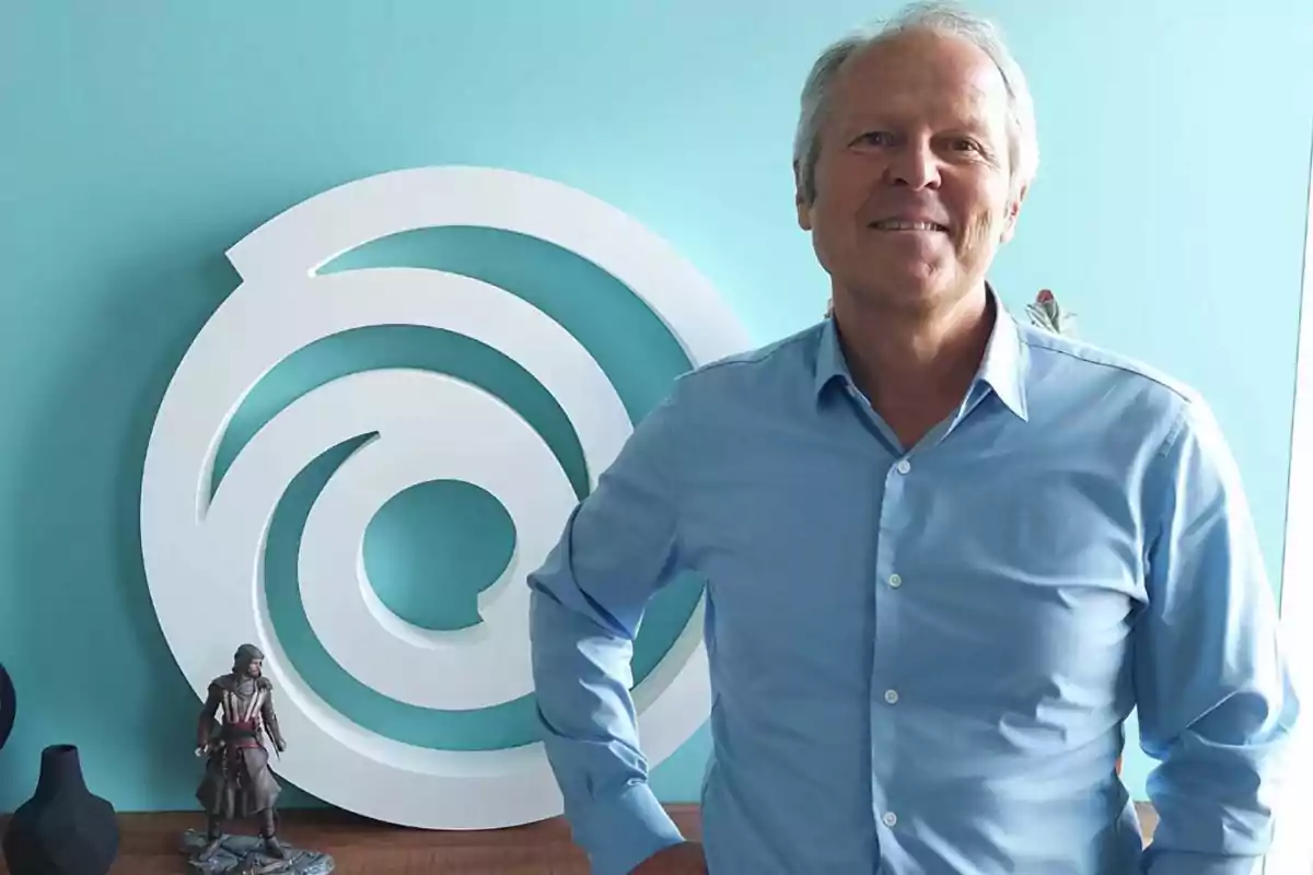 Hombre de cabello canoso con camisa azul claro sonriendo frente a una pared celeste con un gran logo blanco en espiral y una figura de acción sobre una mesa Hombre de cabello canoso con camisa azul claro sonriendo frente a una pared celeste con un gran logo blanco en espiral y una figura de acción sobre una mesa
