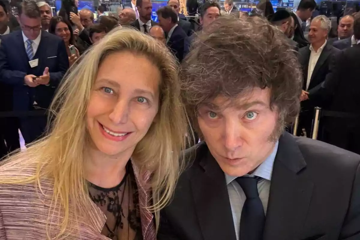 A woman and a man dressed formally pose for a selfie at a crowded event with several people in the background. A woman and a man dressed formally pose for a selfie at a crowded event with several people in the background.