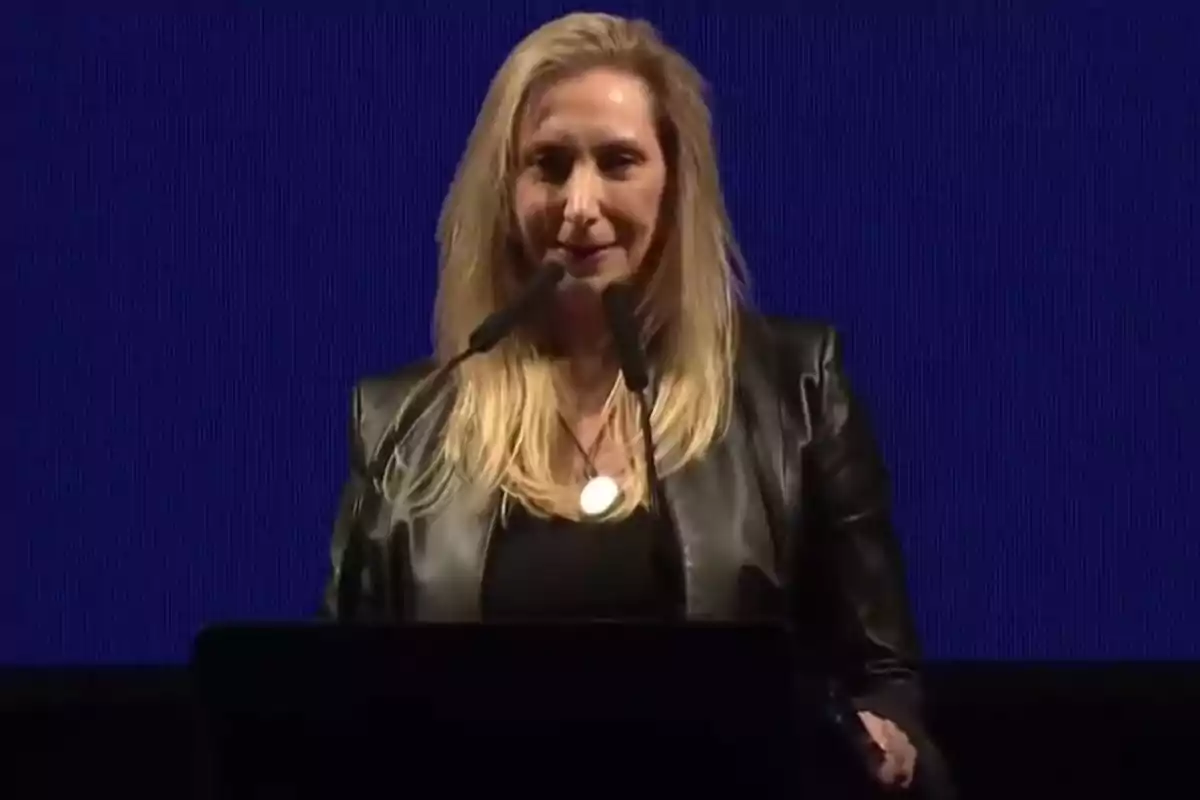 Blonde woman in a black leather jacket speaking in front of two microphones against a blue background