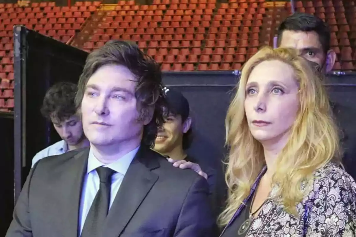 A man in a dark suit and a woman with long blonde hair are together in a place with many empty red seats in the background. A man in a dark suit and a woman with long blonde hair are together in a place with many empty red seats in the background.