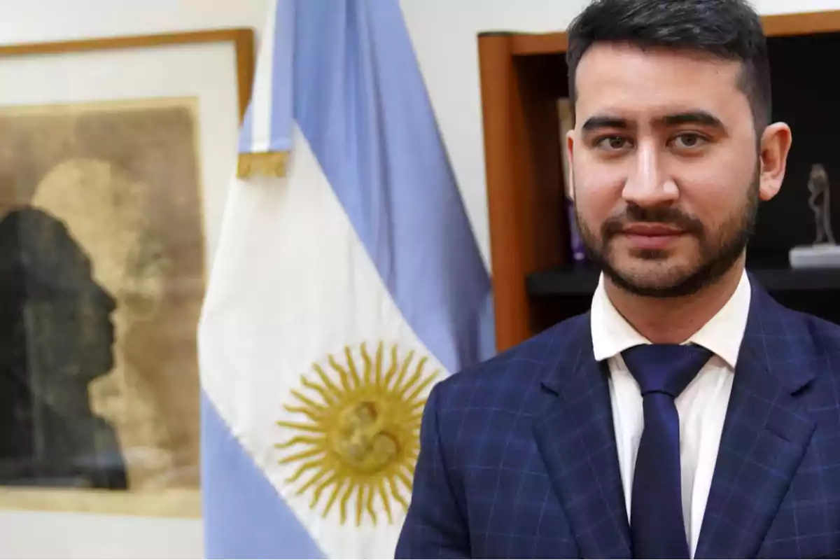 Hombre de traje azul con corbata posando frente a una bandera de Argentina en una oficina