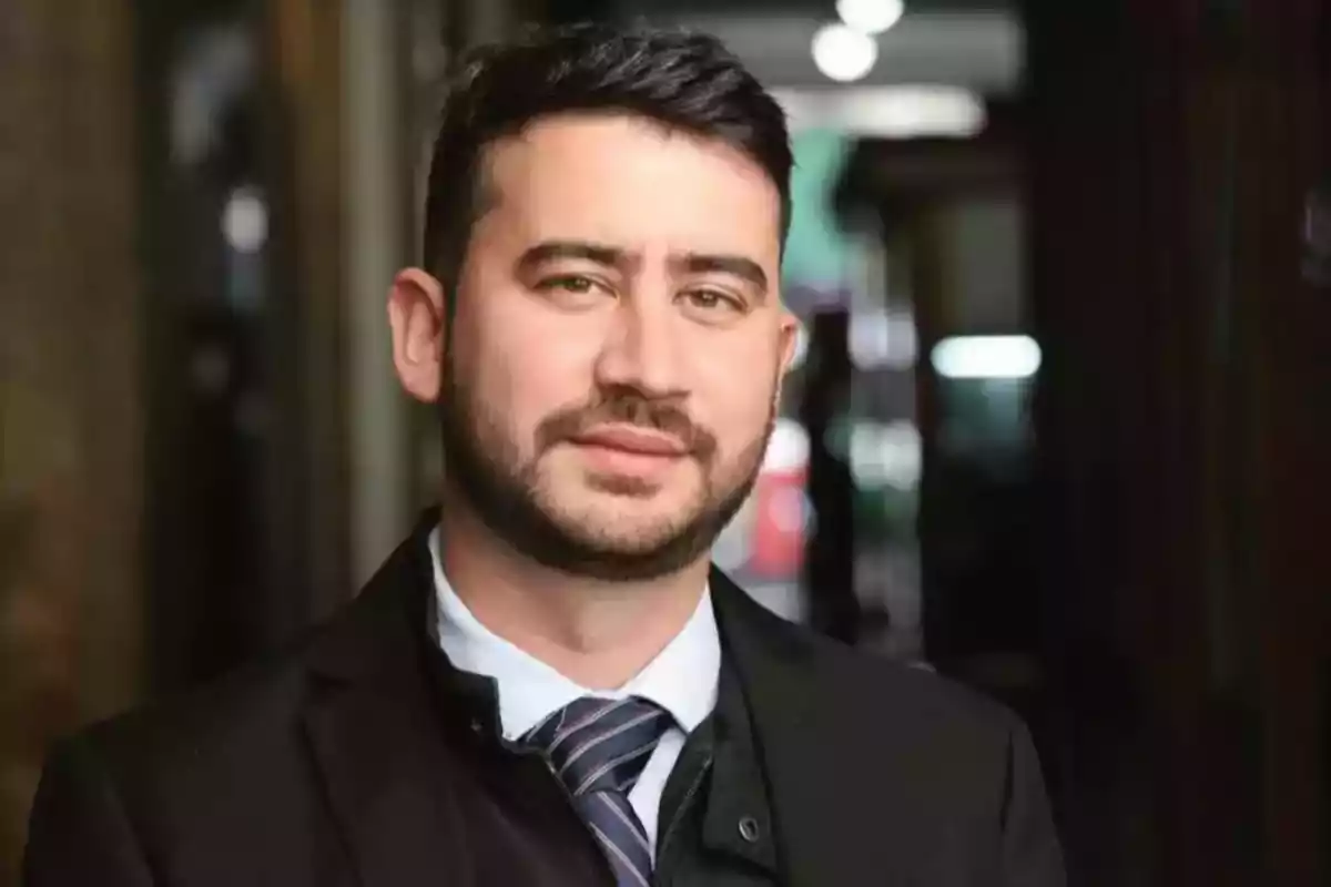 Hombre joven con barba y cabello corto vestido con traje y corbata en un entorno urbano desenfocado