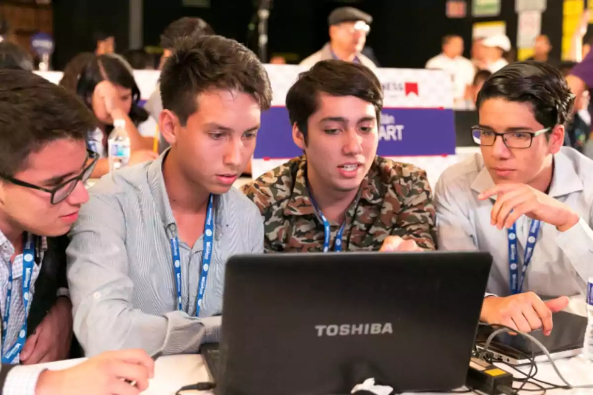 Cuatro jóvenes trabajan juntos frente a una laptop Toshiba en un ambiente de competencia o colaboración académica. Cuatro jóvenes trabajan juntos frente a una laptop Toshiba en un ambiente de competencia o colaboración académica.