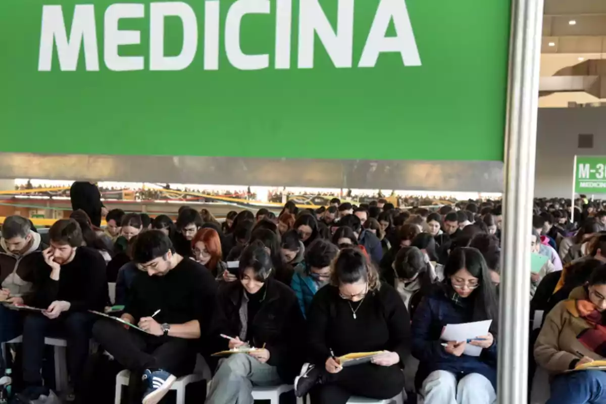 A large group of students sitting under a green sign that says MEDICINE while filling out forms or exams
