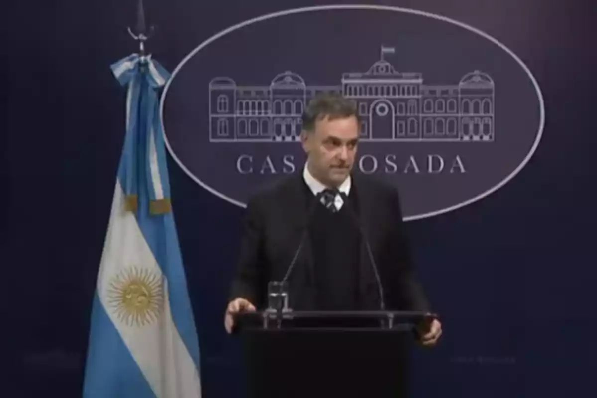 A man in a dark suit speaks at a podium in front of a blue background with an illustration of the Casa Rosada and an Argentine flag beside him. A man in a dark suit speaks at a podium in front of a blue background with an illustration of the Casa Rosada and an Argentine flag beside him.