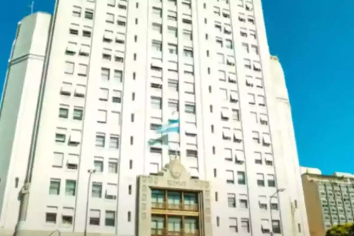Edificio alto de color blanco con muchas ventanas y una bandera argentina ondeando en el centro bajo un cielo despejado