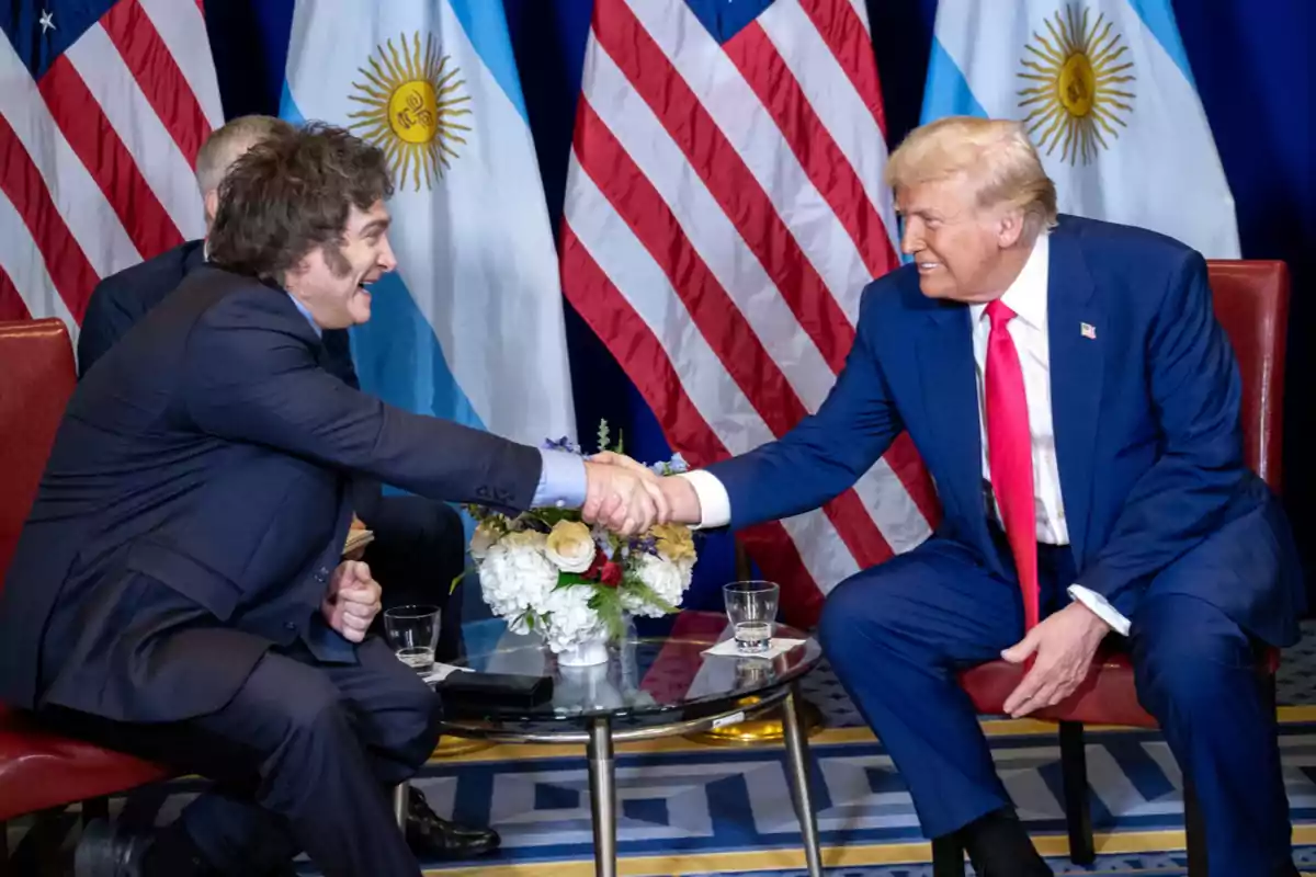 Dos hombres vestidos de traje se dan la mano sentados frente a una mesa de vidrio con flores, al fondo hay banderas de Argentina y Estados Unidos.